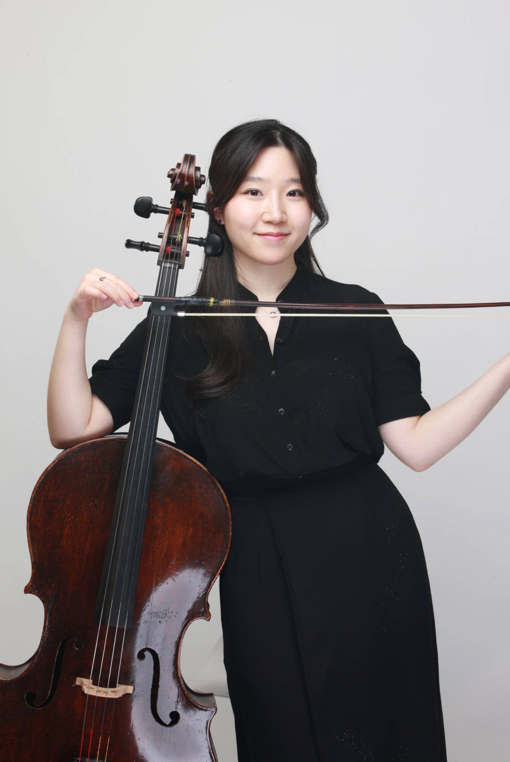 Young In Na in a black shirt poses with a cello, holding the bow horizontally across the instrument's neck against a plain white background.