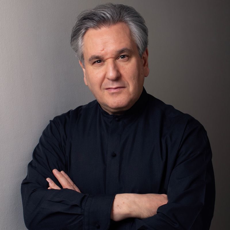 Sir Antonio Pappano, wearing a black top, facing the camera, with his arms folded