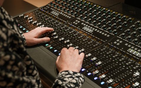 Close-up of hands adjusting faders and knobs on a detailed audio mixing console with illuminated controls.
