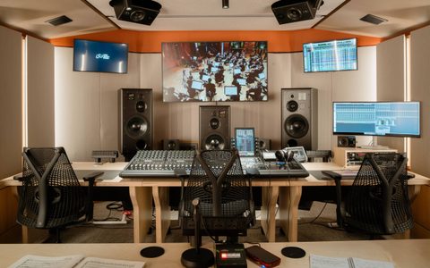 Professional recording studio control room with mixing desks, speakers, and multiple screens, including a large display showing an orchestra session.