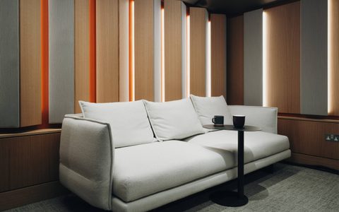 Modern studio lounge with a light-coloured sofa, small round table with mugs, and wooden acoustic wall panels with integrated vertical lighting.