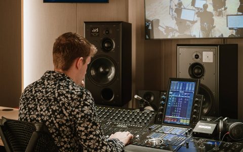 Audio engineer seated at a mixing console, operating controls and a touchscreen display, with studio monitors and a screen showing musicians in the background.