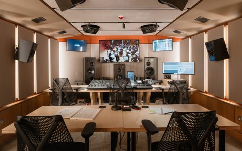 Professional recording studio control room with mixing desks, speakers, and multiple screens, including a large display showing an orchestra session.