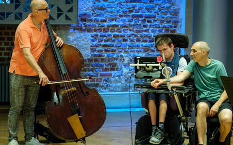 three people making music, one of them on a double bass one of them on a computer with another person sitting next to them.