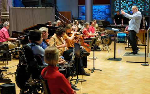 People sitting in a hall making music with different instruments led buy a person who is conducting them.