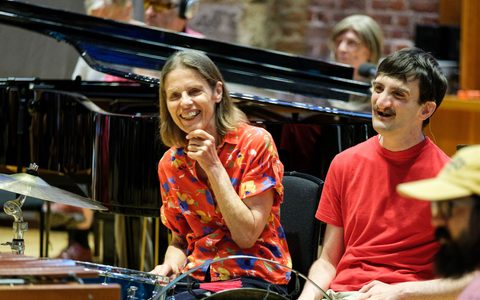 three people participating in a workshop and smiling. Two of them photographed from the front, the third is from the side. They are sitting in front of a piano.